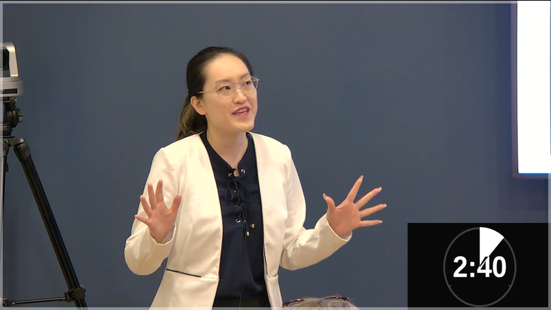 Faculty of Education graduate student and STEAM-3D Maker Lab research assistant Kelly Wang presenting at this year's Three-Minute Thesis competition. In this photo, Kelly is wearing a white blazer over a navy top and is holding her hands out while speaking.